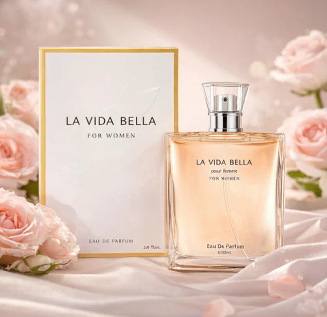 La Vida Bella 100ml Eau de Parfum square glass bottle with a clear cap, standing in front of its elegant white and gold box surrounded by pink roses.